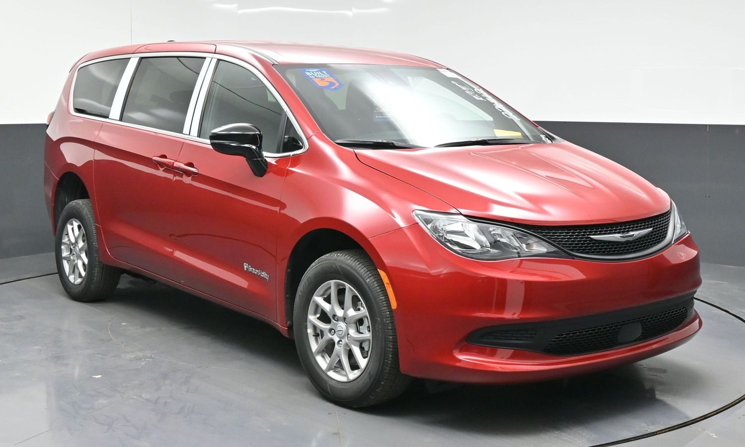 Red minivan (Chrysler Pacifica) parked indoors on a turntable display, facing right with tinted windows and chrome accents.