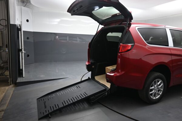 Red minivan with rear hatch open and a folded wheelchair ramp extended to the ground in a clean, indoor testing bay?