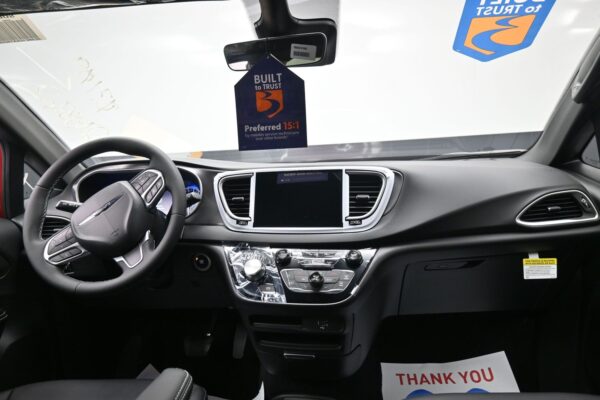 View of a car interior with steering wheel, large center touchscreen, and shiny chrome climate controls; a blue 'Built to Trust' tag hangs from the rearview mirror.
