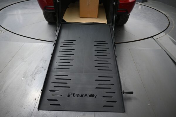 Rear of a red SUV with a BraunAbility wheelchair ramp extended to the ground, inside the cargo area a cardboard box sits on brown packing paper.