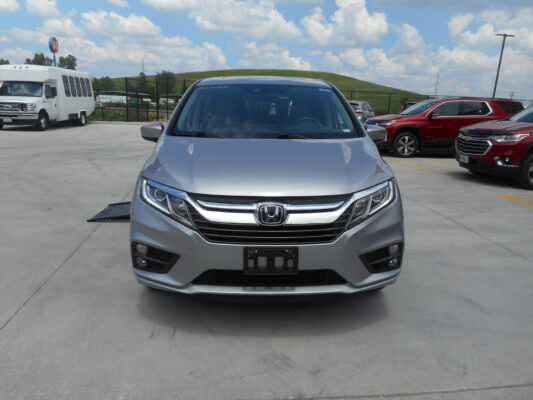 Front view of a silver Honda SUV in a parking lot with other vehicles and a bus in the background under a blue, cloudy sky