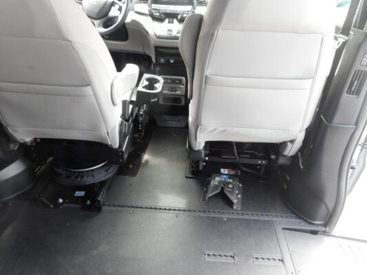 Vehicle interior seen from the back seats; front seats and center console visible with exposed floor hardware and a circular pedestal-style mount on the floor (likely a removable seat/adjustment component).