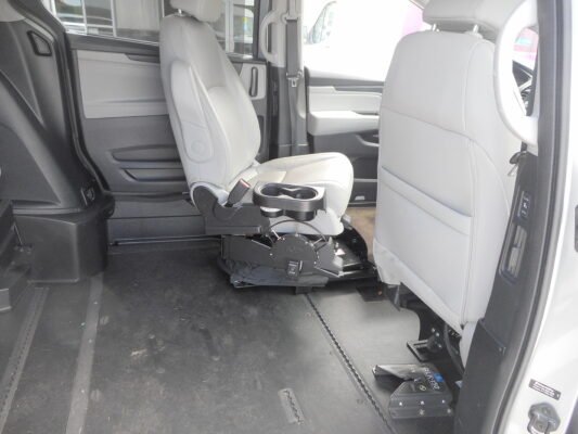 Interior of a vehicle showing a mounted wheelchair lift/seat base and gray passenger seats, with space on the floor for mobility devices