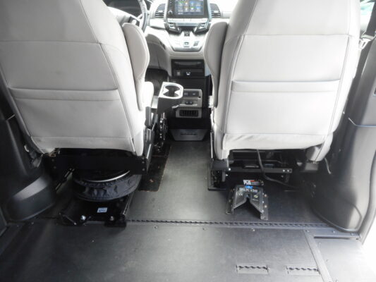 Interior of a vehicle showing the backs of the front seats, center console, and floor-mounted equipment rails or base for a lift/attachment system at the rear area.
