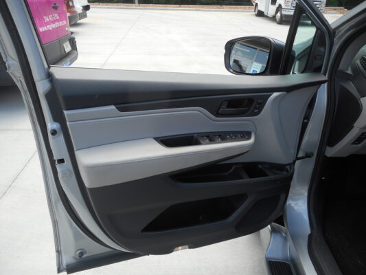 Open car door interior showing gray and black door panel with window controls; outside parking lot visible through the open door and side mirror in view