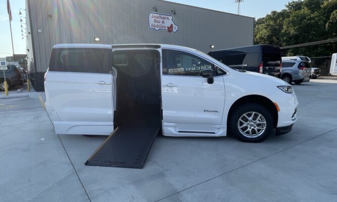 White wheelchair-accessible van with a deployed ramp extending from the side door, parked outside a mobility vehicle shop (BraunAbility branding visible).