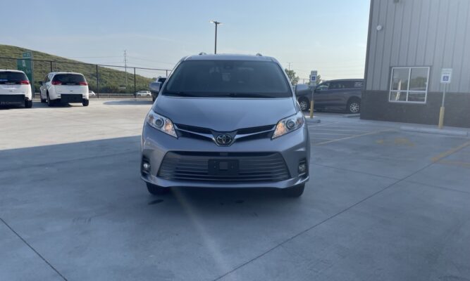 Front view of a silver Toyota minivan facing the camera in a parking lot, with a building and other cars in the background.