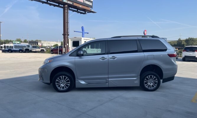Side view of a silver minivan parked in a sunny lot with a tall billboard and other vehicles in the background.