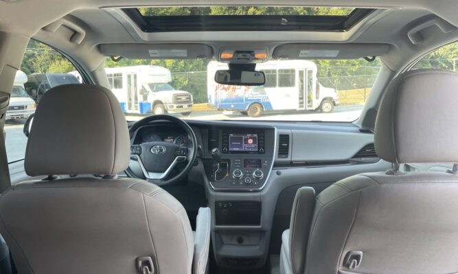Interior of a vehicle viewed from the back seats, showing the dashboard, steering wheel, center touchscreen, and sunroof; buses parked outside the windows.