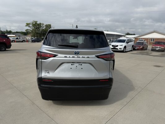 Rear view of a silver Toyota Sienna minivan parked in a lot, with other vehicles and a chain-link fence in the background under a cloudy sky.