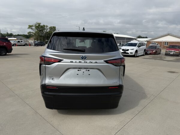 IMG_3377 Rear view of a silver Toyota Sienna minivan parked in a lot, with other vehicles and a chain-link fence in the background under a cloudy sky.