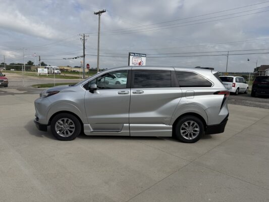 Side view of a silver minivan parked in a dealership lot with a chain-link fence and power lines in the background under a cloudy sky.