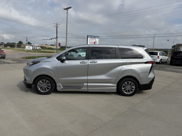 IMG_3378 Side view of a silver minivan parked in a dealership lot with a chain-link fence and power lines in the background under a cloudy sky.