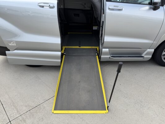 Silver passenger van with a yellow-edged wheelchair ramp extended from the side door, ready for boarding on a paved surface.