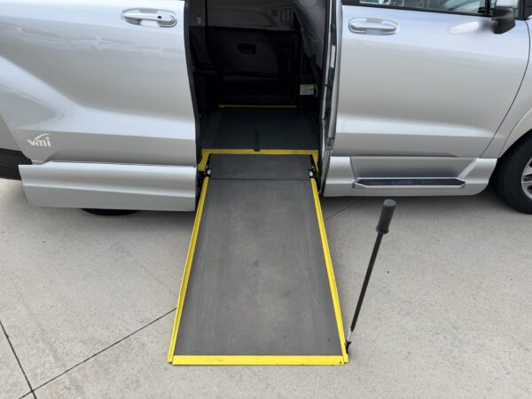 IMG_3382 Silver passenger van with a yellow-edged wheelchair ramp extended from the side door, ready for boarding on a paved surface.