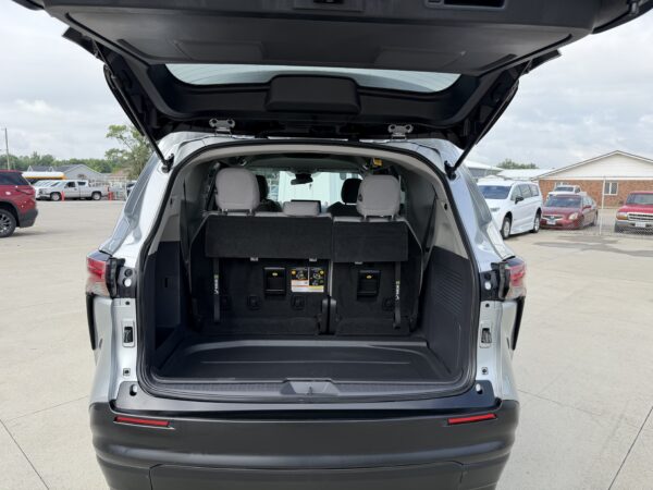 IMG_3385 Open rear hatch of a gray SUV showing folded third-row seats and empty cargo area in a parking lot to the rear showing other cars and a fence in the background.