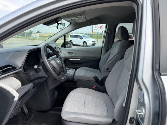 Interior view of a gray SUV showing the driver seat, steering wheel, center console, and front passenger seat with fabric upholstery. Outside through the window is a white van and a chain-link fence.