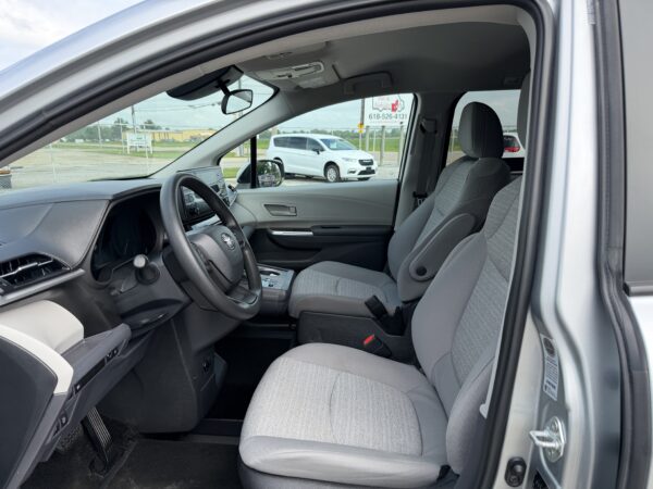 IMG_3386 Interior view of a gray SUV showing the driver seat, steering wheel, center console, and front passenger seat with fabric upholstery. Outside through the window is a white van and a chain-link fence.