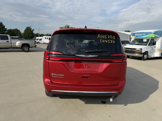 Rear view of a red BraunAbility Pacifica minivan with yellow handwritten numbers on the rear window; a parking lot with trucks and an Americare van in the background.
