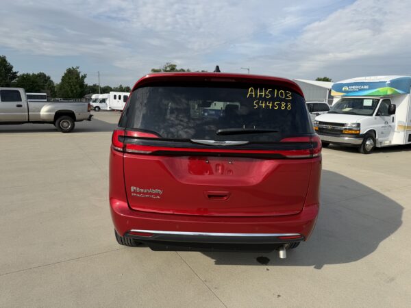 IMG_3470 Rear view of a red BraunAbility Pacifica minivan with yellow handwritten numbers on the rear window; a parking lot with trucks and an Americare van in the background.