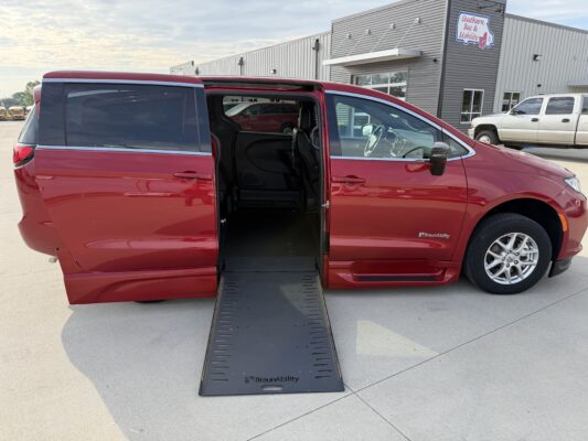 Red wheelchair-accessible van with a lowered ramp extending from side door at a dealership or service center.