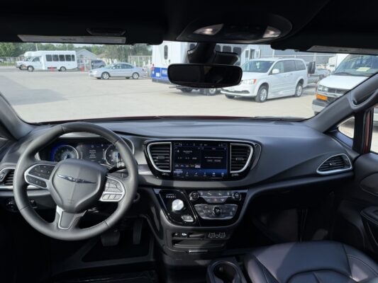 Interior view from the driver’s seat showing the steering wheel, center touchscreen, and rearview mirror, with a parking lot of vans and buses outside.