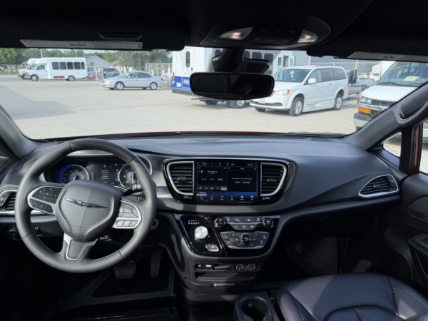 IMG_3478 Interior view from the driver’s seat showing the steering wheel, center touchscreen, and rearview mirror, with a parking lot of vans and buses outside.