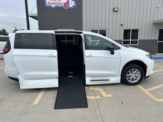 White wheelchair-accessible van with the side door open and a black ramp extended, parked in a handicap space by a gray commercial building.
