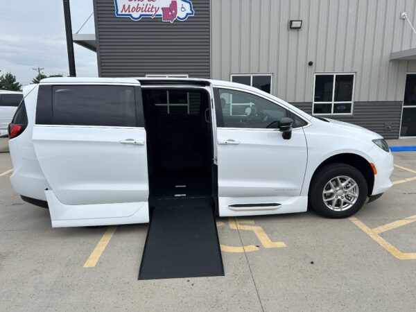 White wheelchair-accessible van with the side door open and a black ramp extended, parked in a handicap space by a gray commercial building.