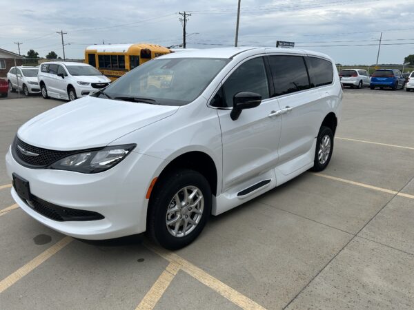 Front three-quarter view of a white Chrysler minivan parked in a lot, with a yellow school bus and other cars in the background.