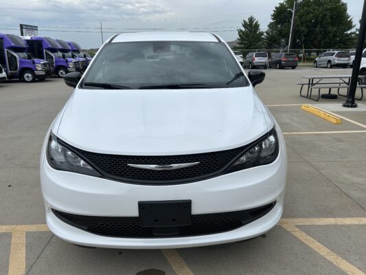 Front view of a white Chrysler minivan parked in a parking lot with purple shuttle buses in the background on the left side.