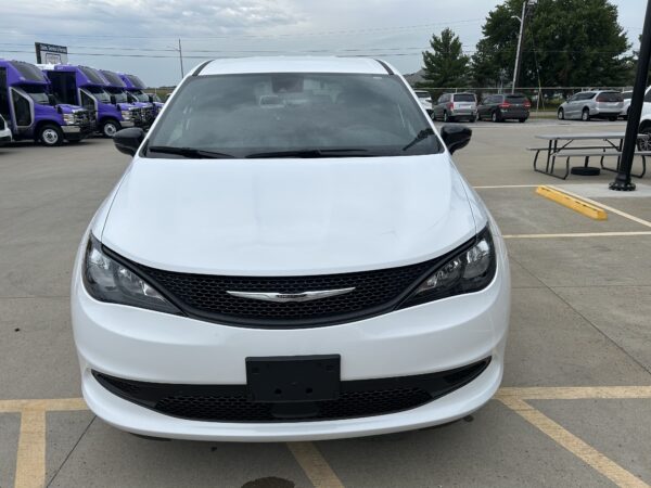 Front view of a white Chrysler minivan parked in a parking lot with purple shuttle buses in the background on the left side.