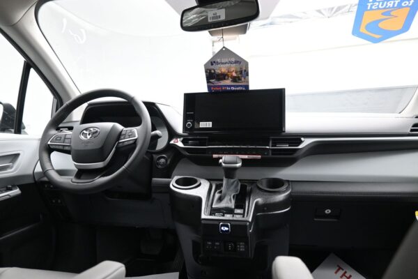Interior of a vehicle cabin featuring a steering wheel, large central touchscreen, gear shifter, and dual cup holders; hanging BraunAbility tag above the dashboard