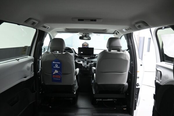 Interior of a white van from the rear, showing two front seats, center console, and a blue BraunAbility Q-STRAINT tag hanging from the driver’s seat.