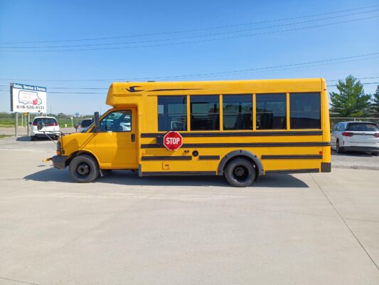 Yellow school bus parked in a lot with its side facing the camera, stop sign visible on the side panel, clear blue sky above.