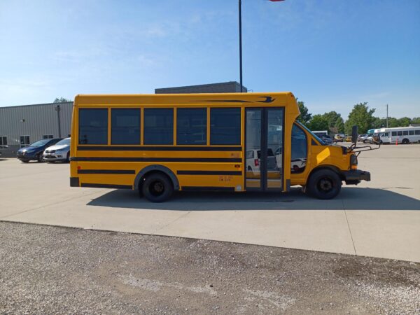 Side view of a yellow school bus parked in a lot with other cars and a metal building in the background, daytime sun.