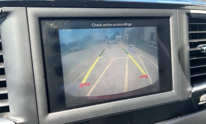 Rearview camera display showing parking guidelines as the car backs up toward a driveway with another car ahead and a building to the right; text reads 'Check entire surroundings'