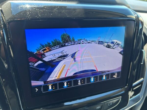 Rear-view camera display showing a parking lot behind the vehicle with guiding lines for safe reversing, including parked cars and a bench visible in the distance.