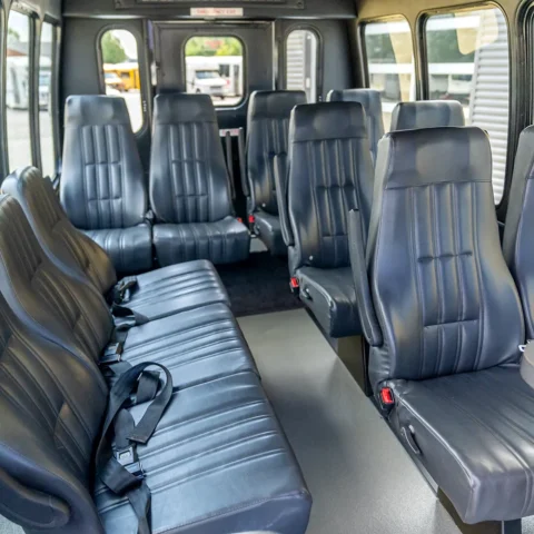 Interior of a passenger shuttle van with multiple rows of black leather seats and a small cupholder table visible at the front/right side.