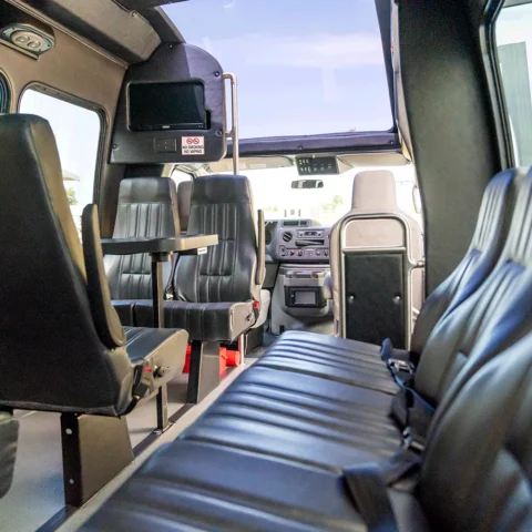 Interior of a passenger minibus showing rows of black leather seats, a central aisle, and a small table, with the driver’s cockpit at the front.