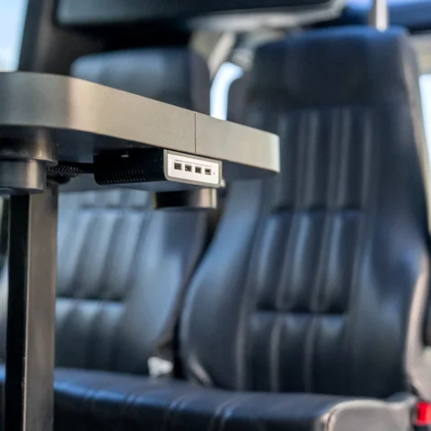 Close-up of a tray table with a small USB port hub attached underneath in a vehicle cabin, blurred seats in the background