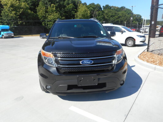 Front view of a black Ford SUV in a parking lot, with a white car beside it and a blue truck in the background, trees and fencing behind.