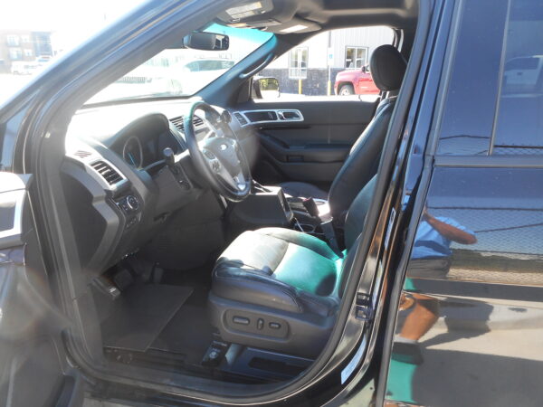 Interior of a dark SUV with the driver’s seat, steering wheel, and center console visible through an open front door; sunlight washes the dashboard. Outside, a parking lot is visible through the window.
