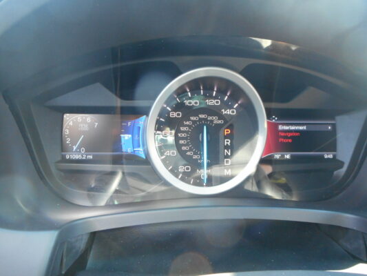 Close-up of a car dashboard: large central speedometer with digital readouts and a left instrument panel showing mileage (91,095.2 mi) and RPMs.
