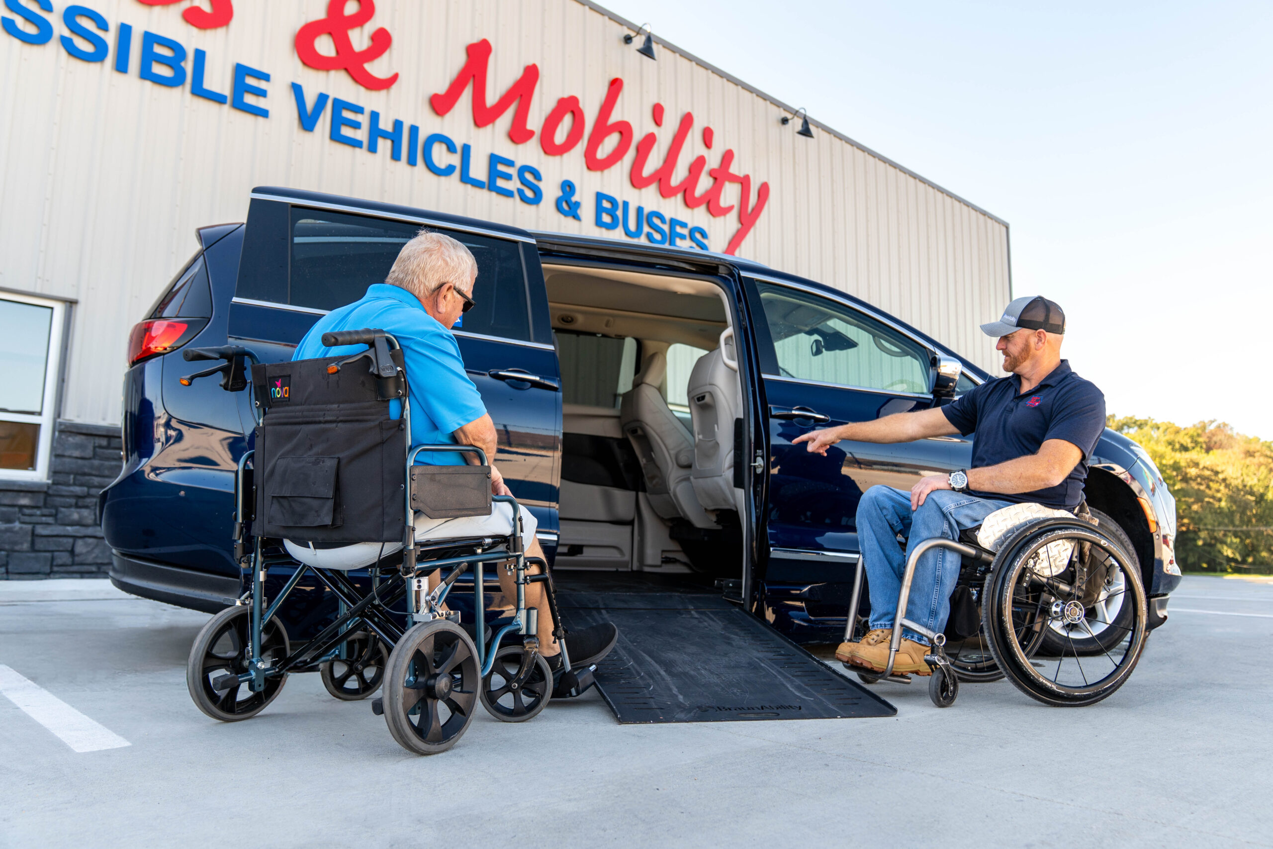 Two men in wheelchairs load into a blue SUV with a side ramp outside a mobility shop.