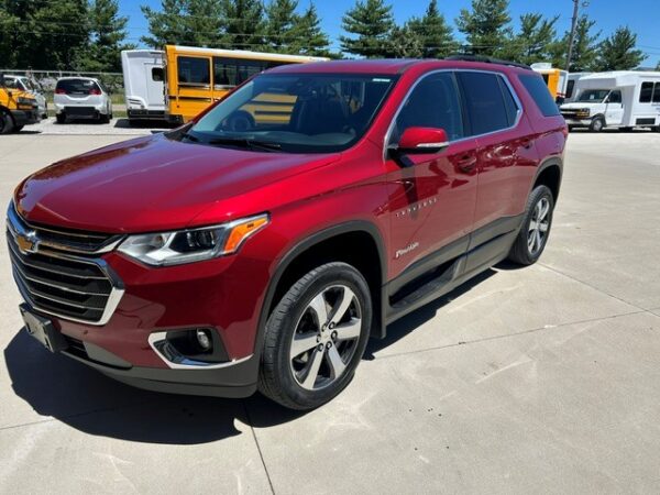 Front three-quarter view of a red Chevrolet Traverse SUV parked in a sunny car dealership lot with yellow buses in the background.