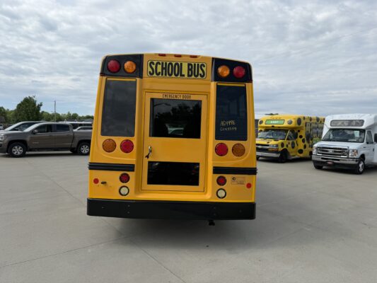 Rear view of a yellow school bus with an 'Emergency Door' label and red/amber taillights, parked in a lot with other buses nearby under a cloudy sky.