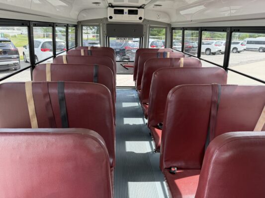 Interior of a school bus with red vinyl seats along a central aisle and large windows on both sides, emergency exit at rear