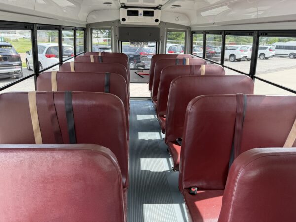 Interior of a school bus with red vinyl seats along a central aisle and large windows on both sides, emergency exit at rear