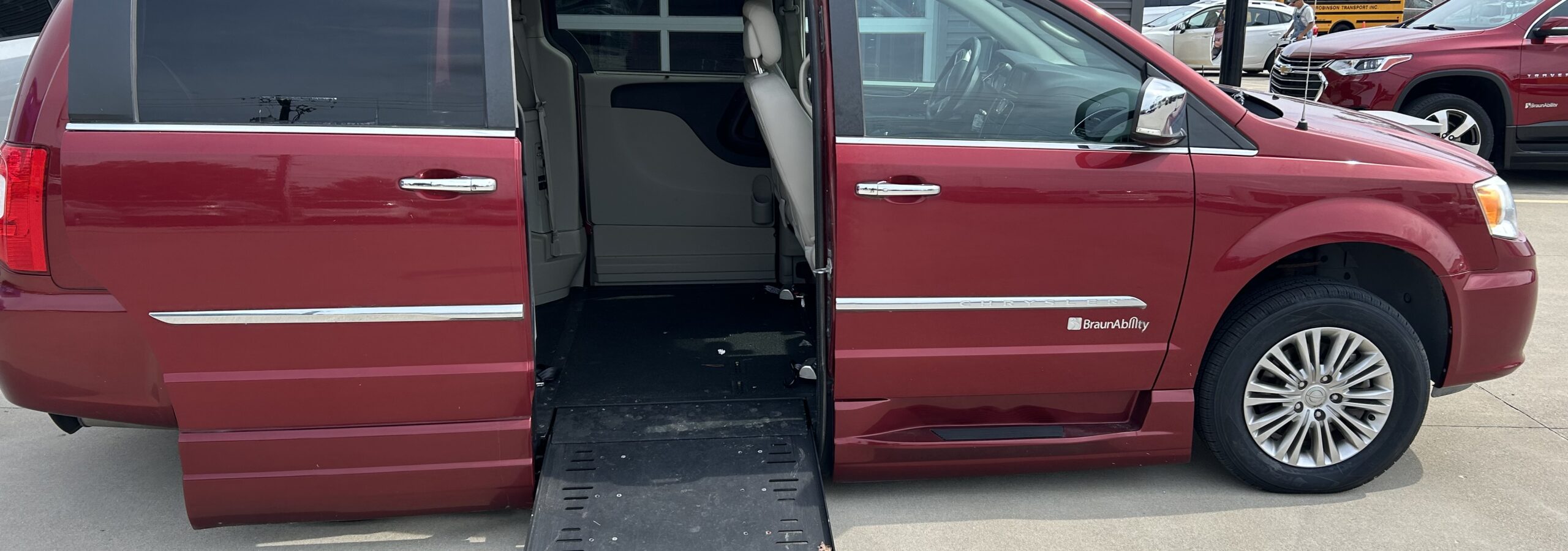Red minivan with a deployed wheelchair ramp extending from the side door, BraunAbility branding visible on the ramp, parked outside a building with other vehicles nearby.